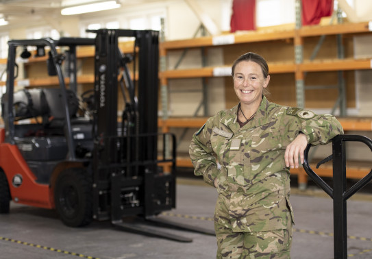Flying Officer Challies-Kolk standing in a warehouse leaning on something. There is a forklift in the background.. 