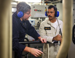 Sub Lieutenant Kamil Rahman (right) talks with another sailor in a plant room, both sailors wearing overalls and blue headphones.