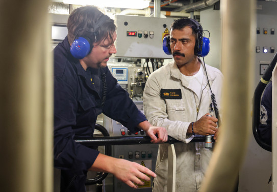 Sub Lieutenant Kamil Rahman (right) talks with another sailor in a plant room, both sailors wearing overalls and blue headphones.
