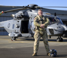 A uniformed aviator stands in front of a NH90 helicopter, looking and smiling at the camera, hands together. 