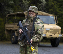 Flying Officer Celine Conaglen stands holding a MARS-L in camouflage uniform in front of a tan-coloured military truck.