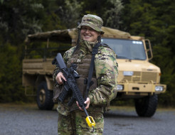 Flying Officer Celine Conaglen stands holding a MARS-L in camouflage uniform in front of a tan-coloured military truck.