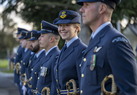 A uniformed RNZAF aviator stands in a line of other uniformed aviators - they are looking at the camera and smiling. The others are looking to the left of the camera and are out of focus. 