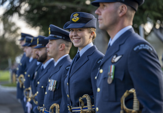 A uniformed RNZAF aviator stands in a line of other uniformed aviators - they are looking at the camera and smiling. The others are looking to the left of the camera and are out of focus. 