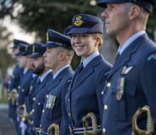 A uniformed RNZAF aviator stands in a line of other uniformed aviators - they are looking at the camera and smiling. The others are looking to the left of the camera and are out of focus. 