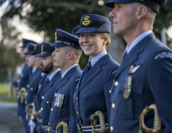 A uniformed RNZAF aviator stands in a line of other uniformed aviators - they are looking at the camera and smiling. The others are looking to the left of the camera and are out of focus. 