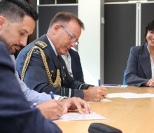 The Chief of Defence Force sits at a table signing a piece of paper alongside another male who also signs a piece of paper. Around the table are people wearing various business attire as they watch on.