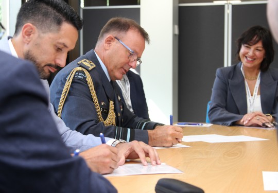 The Chief of Defence Force sits at a table signing a piece of paper alongside another male who also signs a piece of paper. Around the table are people wearing various business attire as they watch on.
