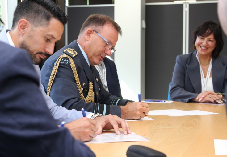 The Chief of Defence Force sits at a table signing a piece of paper alongside another male who also signs a piece of paper. Around the table are people wearing various business attire as they watch on.
