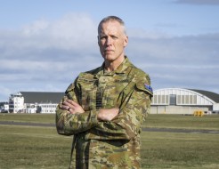 A male in an australian camoflauge military uniform stands arms folded with an airfield and hangars behind him. He has an Australian flag on his arm and shows the rank of Group Captain on his rank slide.