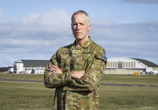 A male in an australian camoflauge military uniform stands arms folded with an airfield and hangars behind him. He has an Australian flag on his arm and shows the rank of Group Captain on his rank slide.