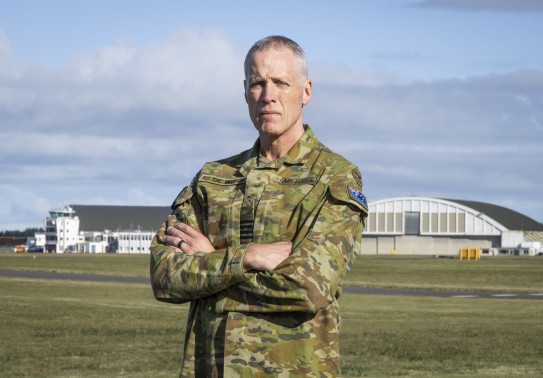 A male in an australian camoflauge military uniform stands arms folded with an airfield and hangars behind him. He has an Australian flag on his arm and shows the rank of Group Captain on his rank slide.
