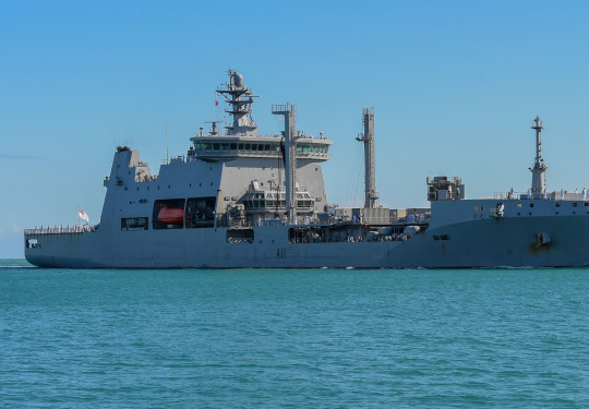 Large grey tanker ship with orange life boats slowly passes land as it arrives into Pearl Harbour.