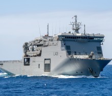 A large grey ship sails through blue water under clear skies,