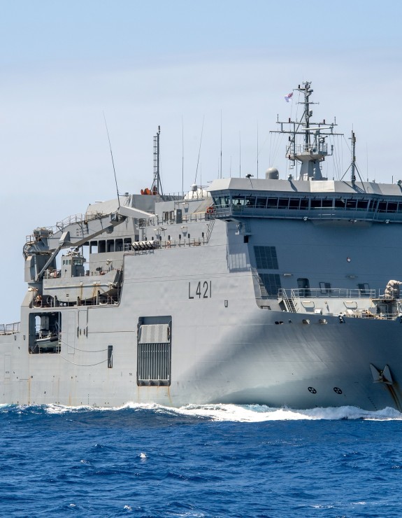 A large grey ship sails through blue water under clear skies,