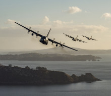 Three C-130H aircraft flying formation in a line over a harbour. All three aircraft are banking in a turn.