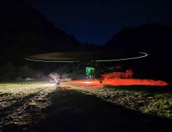 A large grey helicopter has it's rotor blades spinning during night time hours whilst stationery on a field.