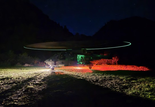 A large grey helicopter has it's rotor blades spinning during night time hours whilst stationery on a field.