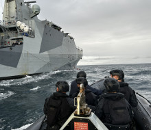 A RHIB with personnel from HMNZS Matataua wearing black uniform and helmets moves over the ocean along side HMS Spey, a grey Royal Navy ship.