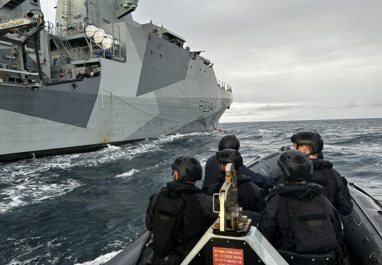 A RHIB with personnel from HMNZS Matataua wearing black uniform and helmets moves over the ocean along side HMS Spey, a grey Royal Navy ship.