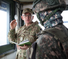Two soldiers in different military uniform talk in a small room with large windows.