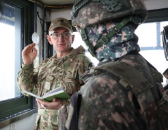 Two soldiers in different military uniform talk in a small room with large windows.
