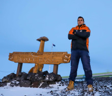 LCPL Darryl Spencer stands next to the wooden carved 'Scott Base' sign. He is wearing an Antartica New Zealand jacket which is bright orange and black, his arms are crossed and his tan boots have snow on the ends.