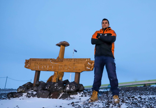 LCPL Darryl Spencer stands next to the wooden carved 'Scott Base' sign. He is wearing an Antartica New Zealand jacket which is bright orange and black, his arms are crossed and his tan boots have snow on the ends.