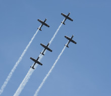 Five small planes fly in a cross formation while releasing smoke as a part of a flight display.