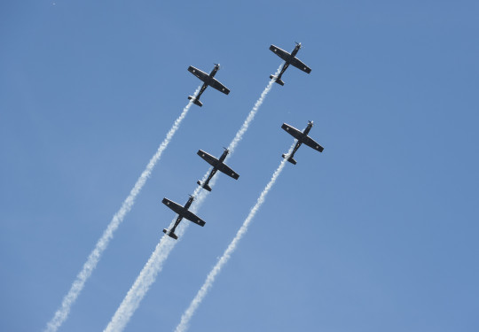 Five small planes fly in a cross formation while releasing smoke as a part of a flight display.