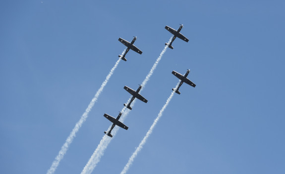 Five small planes fly in a cross formation while releasing smoke as a part of a flight display.