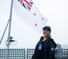 A woman dressed in a dark blue rain jacket and cap stands in front of a Navy Flag on board a Navy Ship.