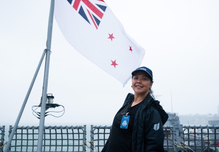 A woman dressed in a dark blue rain jacket and cap stands in front of a Navy Flag on board a Navy Ship.