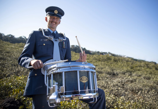 Percussionist Corporal Jeremy Richardson stands with a drum, all in uniform with greenery and sky in the background. 