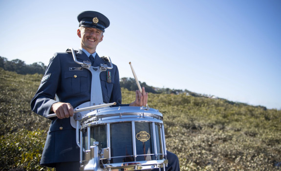Percussionist Corporal Jeremy Richardson stands with a drum, all in uniform with greenery and sky in the background. 