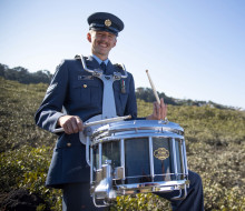 Percussionist Corporal Jeremy Richardson stands with a drum, all in uniform with greenery and sky in the background.