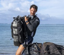 A diver holds a dive tank while in a dive t-shirt and shorts with a pair of goggles around his neck.