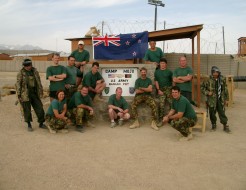 A group of soldiers dressed down wearing green t-shirts and camouflage trousers pose underneath a sign saying 'Camp Mojo'. The sign is on a hut, the New Zealand flag is positioned above and we see fencing, barbed wire and sand in the surrounding environme