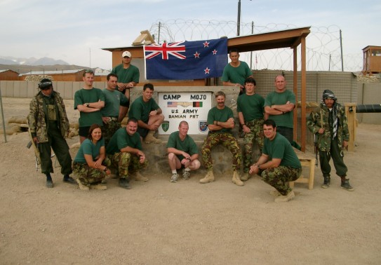 A group of soldiers dressed down wearing green t-shirts and camouflage trousers pose underneath a sign saying 'Camp Mojo'. The sign is on a hut, the New Zealand flag is positioned above and we see fencing, barbed wire and sand in the surrounding environme