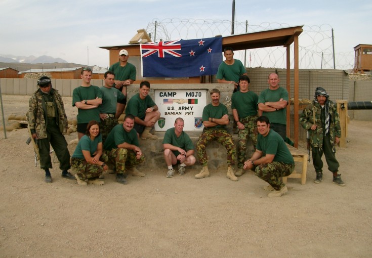 A group of soldiers dressed down wearing green t-shirts and camouflage trousers pose underneath a sign saying 'Camp Mojo'. The sign is on a hut, the New Zealand flag is positioned above and we see fencing, barbed wire and sand in the surrounding environme
