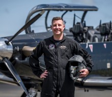 A military pilot smiles broadly whilst wearing a black flying suit. He stands in front of a small black aircraft with one hand on his hip and the other holding his helmet. 