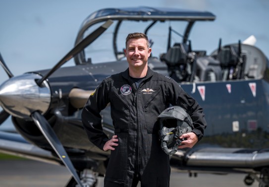 A military pilot smiles broadly whilst wearing a black flying suit. He stands in front of a small black aircraft with one hand on his hip and the other holding his helmet. 