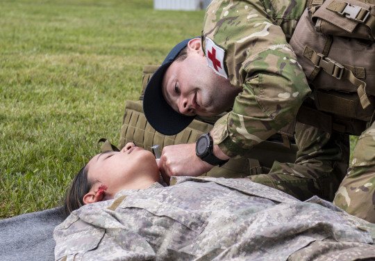 Leading Aircraftman Scott Endres wearing a baseball cap, camouflage uniform with a red cross on his left arm, tends to a person on the ground in a field.