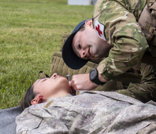 Leading Aircraftman Scott Endres wearing a baseball cap, camouflage uniform with a red cross on his left arm, tends to a person on the ground in a field.