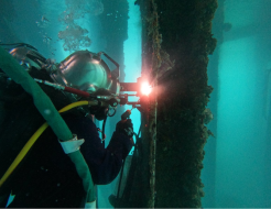 A diver undertakes a welding task in turquoise water. The diver is in a diving helmet and full diving and safety apparatus.