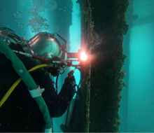A diver undertakes a welding task in turquoise water. The diver is in a diving helmet and full diving and safety apparatus.