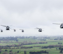 Seven of the Royal New Zealand Air Force’s NH90 helicopters fly in the sky over green land. The sky is very cloudy and grey. The image of the aircraft is taken from behind so the perspective is the back of al the aircraft.
