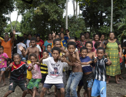 Children in Papua New Guinea pose for the camera while their families are in the background by trees.