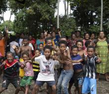 Children in Papua New Guinea pose for the camera while their families are in the background by trees.