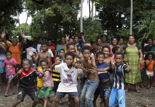 Children in Papua New Guinea pose for the camera while their families are in the background by trees.
